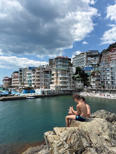 The image depicts a coastal scene in Kiparisnoe, Ukraine, characterized by a dense collection of multi-story buildings lining a waterfront. In the foreground, two young boys are seated on large rocks, facing away from the camera and towards the water. One boy is wearing dark blue swim trunks, and the other is out of focus behind him. The water is a vibrant turquoise color, and the shoreline is lined with a pebbly beach populated by many people sunbathing and swimming. A small harbor area with several boats is visible to the left, and further along the coast, more buildings are clustered on the hillside. The sky is partly cloudy with dramatic, dark clouds interspersed with patches of blue.