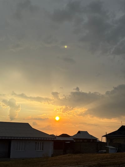 This image captures a sunset over a rural or semi-rural setting in Baş-Bulak, Kyrgyzstan. The sun, a bright orange orb, is low in the sky, casting a warm glow across the clouds. The sky is a mix of dramatic, dark storm clouds on the left and lighter, illuminated clouds on the right, suggesting a transition in weather or a dramatic atmospheric effect.

Several buildings are visible, characterized by their angular roofs and brick construction, typical of residential or small industrial structures in the region. One building, in the foreground, has a light-colored facade with horizontal siding and multiple windows. Another building behind it is made of dark brick. The presence of these structures, along with the patchy, dry grass on the right side of the frame, indicates human habitation and land use.

The time of day is clearly sunset, given the low angle of the sun and the warm, golden light. The weather appears to be dynamic, with both heavy cloud cover and illuminated areas, potentially indicating the aftermath of a storm or the approach of evening. The overall mood is one of natural beauty and quiet observation, with the striking sky dominating the scene. There is no visible text in the image.
