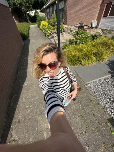 A selfie taken at street level in Dronten, Netherlands. A woman with sunglasses and a striped shirt is holding the camera. A handbag is visible. She is sticking out her tongue playfully. Behind her is a sidewalk with vegetation and buildings on either side. A house with a brick wall is on the lower left, and a house with a grey wall and air conditioning unit is on the upper left. Gravel and bushes are also visible on the upper right. The scene appears to be taken during daylight under sunny conditions.