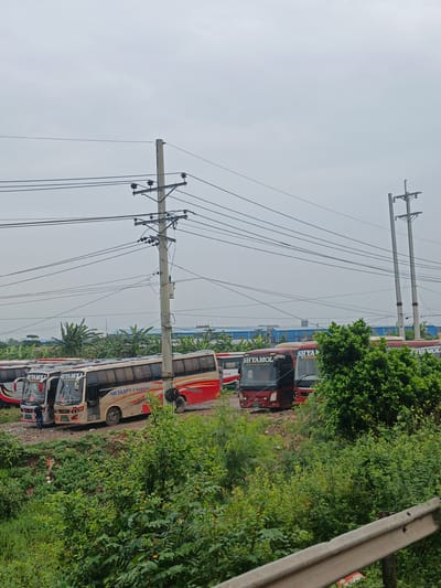 The image depicts a street scene in Boliarpur, Bangladesh, featuring a row of parked buses under an overcast sky. The buses, varying in color from red and cream to dark red, are positioned on a paved area. Some buses display the brand name "SHYAMOLI PARIBAHAN." Power lines crisscross the sky, supported by concrete utility poles. Lush green vegetation, including trees and bushes, lines the foreground and frames the scene. A silver guardrail is visible in the lower right corner. The background reveals more greenery and industrial-looking buildings. A person can be seen standing near the entrance of one of the buses.