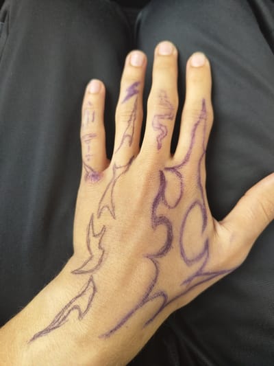 The image shows a close-up of a person's left hand with purple marker drawings on it. The drawings appear to be of flames or tribal designs. The hand is resting on a dark fabric, possibly clothing. There are no visible identifying landmarks or details suggesting a specific location in Hadzhidimitrovo, Bulgaria.