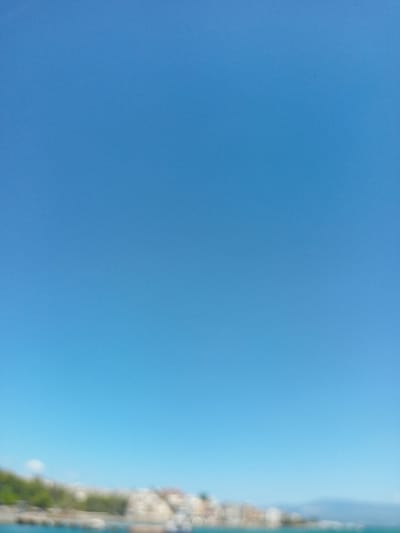 The image shows a bright blue sky dominating the frame. The bottom portion of the image depicts a blurred coastal landscape, likely the city of Chalkida, Greece. A line of indistinct buildings, some with reddish-brown roofs, are visible along the coastline, interspersed with greenery. The water is also visible and appears turquoise. The scene suggests a sunny day.