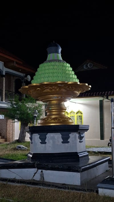 The image depicts a night-time outdoor scene in Lhokdagang, Indonesia, focusing on a decorative monument. The monument, positioned centrally, is a multi-tiered structure. The base is a dark, stepped pedestal, the lowest tier of which is cracked. Above this is a grey layer with dark grey decorative elements resembling small columns. The main body of the monument is a large, ornate, golden urn with intricate carvings. Capping the urn is a conical structure covered in bright green, scale-like decorative elements, topped with a dark blue dome.

In the background, to the left, is a building with a red-tiled roof and a balcony. To the right, another building with white tiled walls and yellow-framed windows is visible. A tree with sparse foliage stands to the left of the monument. The ground is covered with dry grass. The overall lighting suggests artificial illumination from the monument and surrounding buildings. No people are present in the frame, and no specific actions or interactions are depicted. The scene is static and appears to be a photographic record of the monument.