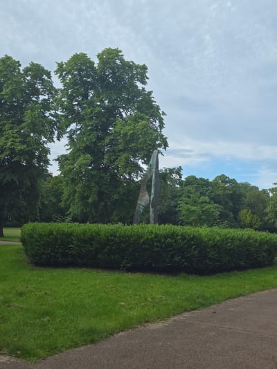 The image depicts a cemetery with a paved path, a meticulously maintained green lawn, and a tall, dense hedge. In the background, lush green trees form a dense canopy. A distinctive bronze sculpture, with a weathered patina, stands prominently among the trees. The sky above is a soft blue with wispy white clouds, suggesting it's a clear day, likely in the late morning or early afternoon. The scene is tranquil and serene, evoking a sense of peace. The overall impression is one of a well-kept and beautiful resting place, harmoniously blending natural elements with artistic expression. The image appears to be taken in Stockton-on-Tees, United Kingdom, given the location context. There is no visible text in the image.