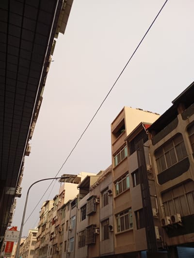 The image captures a street in Kaohsiung, Taiwan, looking upwards at the buildings on either side. The buildings are of mixed architectural styles, predominantly multi-story residential structures. Most units are equipped with air conditioning units mounted on the exterior walls. A street lamp arches into the frame from the lower left. A single black wire stretches diagonally across the sky. The sky appears overcast. Signage is visible at the bottom left of the frame with Chinese text, one is for a traditional Chinese medicine clinic. The perspective suggests the shot was taken from street level. No people are clearly visible.