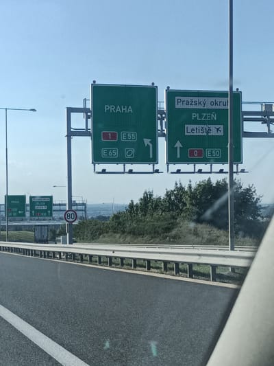 This media file captures a road scene in Kuří, Czechia, likely during the daytime under clear skies. The primary focus is on a series of green highway signs indicating directions and route numbers. On the left, a smaller sign reads "PRAHA" with an arrow pointing left, and below it, "Pražský okruh" and "Plzeň" with arrows indicating straight ahead.
To the right, a larger, more prominent sign displays "PRAHA" at the top. Below this, it shows "Pražský okrul" and "PLZEN" with an airplane icon next to "Letiště" (which translates to airport). The sign further indicates route information: "1 E55" for a lane going straight, "E 65" for another lane going straight, and "0 E50" for a lane going straight with an upward arrow.
In the foreground, a paved highway with white lane markings is visible, along with a metal guardrail. Further in the background, there are trees and a distant, hazy landscape, suggesting a rural or semi-urban setting. A speed limit sign with "80" is also visible. The overall scene conveys information and navigation for drivers on a highway. There are no people visible in the frame.