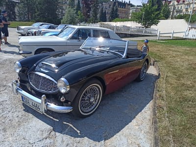 The image captures an outdoor event, likely a car show, in the city of Mariánské Lázně, Czechia. In the foreground, a black convertible classic car with a red lower body is prominently displayed. Its chrome grille, headlights, and wire-spoke wheels are gleaming under the sun. The car's license plate reads "5Z4 4464". Behind it, a row of other vintage vehicles, including a light-colored sedan and what appears to be a white Rolls-Royce, are parked. To the right of the black convertible, a young boy wearing a blue baseball cap and a striped shirt stands near a grassy area. In the background, a white picket fence lines a pathway, and ornate, multi-story buildings with distinct architectural features are visible, characteristic of Mariánské Lázně's spa town aesthetic. The scene is bathed in bright daylight, with long shadows cast on the paved ground.