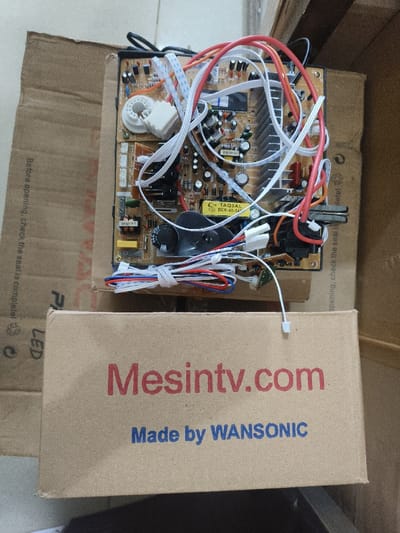 The image displays an electronic circuit board, consistent with a television mainboard or power supply, resting on a partially open brown cardboard box in Alue Mudem, Indonesia. In the foreground, a closed brown cardboard box prominently features "Mesintv.com" in red text and "Made by WANSONIC" in blue text. The circuit board, centrally positioned, is populated with various electronic components including multiple capacitors, resistors, an aluminum heatsink, and a transformer labeled "TAQJAL BCK-40-14". A complex network of multi-colored wires (red, white, blue, orange, and multi-conductor ribbon cables) interconnects different sections of the board; a bundle of these wires is secured with a twist tie. A single white connector hangs freely from the board, extending towards the foreground box. Behind the circuit board, a second, larger brown cardboard box is visible at an angle, displaying partial text on its side such as "Before opening, check the seal is complete!" and "LED," along with international recycling symbols. The visible floor surface beneath the boxes is light-colored. No human subjects are present in the scene.
