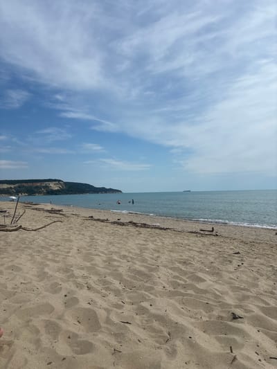The image depicts a serene beach scene in Bliznatsi, Bulgaria. The foreground is dominated by sandy terrain with scattered driftwood. In the mid-ground, the gentle waves of the Black Sea lap the shore, and several individuals are visible in the water, some swimming and others wading. Beyond the water, a long, tree-covered cliff face stretches across the horizon, with a small ship visible in the distance on the calm sea. The sky is a clear blue, with wispy white clouds scattered across it.