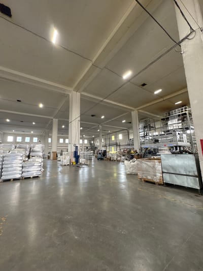 The image depicts the interior of a large warehouse. The warehouse has a high ceiling with exposed beams and fluorescent lighting. Large industrial machinery, possibly for manufacturing, is visible in the background on the right side of the image. Stacks of goods, wrapped in plastic, are organized on pallets in the foreground and mid-ground, suggesting a storage and distribution facility. The floor is a concrete surface. The overall impression is of an active industrial or logistical space.
