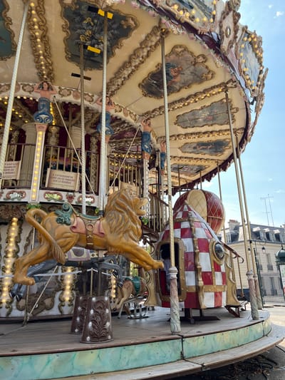 The image shows a close-up, low-angle view of a carousel in Rambouillet, France. The carousel features ornate painted panels and is decorated with lights. A yellow lion figure is prominently displayed in the foreground, along with a rocket-shaped ride. The background shows buildings and a clear blue sky. Signs in French indicate rules for carousel riders, such as "IL EST INTERDIT DE BOIRE OU DE MANGER SUR LE MANÈGE" (It is forbidden to drink or eat on the carousel). The carousel appears to be stationary and no people are clearly visible on the ride.