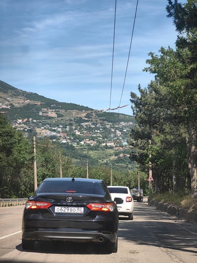 The image shows a view from a vehicle traveling on a road with a mountainous landscape on the left and a dense forest on the right. The road is paved and appears to be a two-lane road with a guardrail on the left side. Several cars are visible on the road, including a black Toyota Camry in the foreground, a white car behind it, and a utility vehicle further ahead. The mountainous area is dotted with numerous houses and buildings, suggesting a settlement. Power lines are strung across the sky, supported by utility poles. The weather appears to be clear and sunny, with a blue sky and some scattered clouds. The scene is consistent with a rural or suburban area near a coast or mountains, likely in the region of Kiparisnoe, Ukraine, given the context.