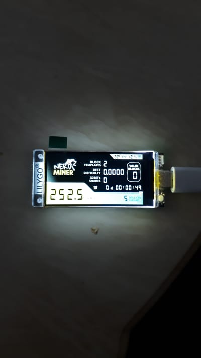 The image shows a close-up of a small electronic device, likely a cryptocurrency miner, with a black and white screen. The device is illuminated by an external light source, casting a glow around it. The screen displays various statistics, including "BLOCK TEMPLATES 2", "BEST DIFFICULTY 0.0000", "32BITS SHARES 0", "VALID BLOCKS 0", and "5 MILLION HASHES". It also shows a timestamp "32 14:12" and a timer "0:00:00:49". The brand name "NERD MINER" is prominently displayed on the screen, along with the "LILYGO" logo at the bottom. The device appears to be connected to a USB cable at the top. The background is dark and textured, suggesting it is placed on a surface in a dimly lit environment. There are no people visible in the image. The overall impression is of a functional piece of technology in operation, possibly in a workshop or personal computing space.