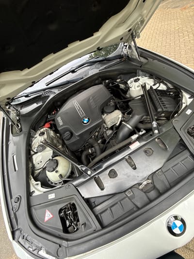 The image shows the engine bay of a white BMW vehicle. The hood is open, revealing the engine, various hoses, and other engine components. A black plastic engine cover with "BMW TwinPower Turbo" and the BMW logo is prominently visible. The car is parked on a paved surface, likely a driveway or street, with a brick-like pattern. The background shows a blurry view of trees and a building, suggesting an outdoor urban or suburban setting. The overall impression is that of a car parked outdoors, possibly for inspection or maintenance. The location is Amstelveen, Netherlands.