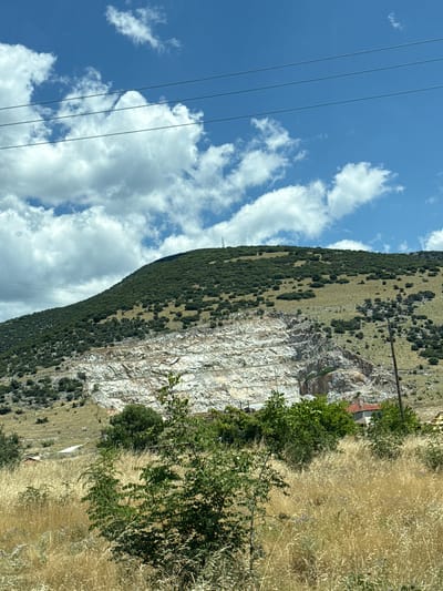 The image depicts a mountainous landscape in Prosotsani Municipal Unit, Greece, characterized by a large, exposed rock face on the side of a densely vegetated hill. The rock face exhibits distinct horizontal striations, suggesting a quarry or a natural geological formation. The upper portion of the hill is covered in a thick layer of dark green vegetation, interspersed with patches of dry, yellowish grass. The foreground is dominated by tall, dry grasses and some leafy green bushes, indicating a natural, possibly semi-arid environment. In the middle ground, several small structures with red roofs are visible amidst the greenery, hinting at human habitation. Power lines are strung across the bright blue sky, which is partially filled with fluffy white clouds. The overall scene conveys a sense of rugged natural beauty, with elements of human presence integrated into the landscape.