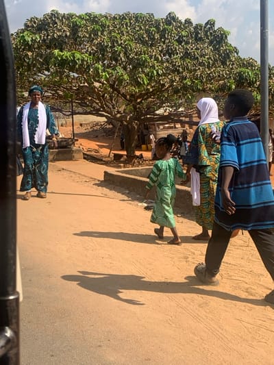 This image captures an outdoor scene in Egbedi, Nigeria, under a bright sky with scattered clouds. The ground is an unpaved, light brown dirt surface, with visible shadows indicating strong daylight.
Four primary individuals are visible. In the mid-ground left, an adult female stands facing the camera. She wears a teal-patterned long dress with a matching headwrap and a white scarf draped over her shoulders. Her footwear consists of light-colored open-toed sandals. In the mid-ground center, a young girl walks away from the camera, wearing a long green dress patterned with white concentric spirals. Her hair is styled in braids. Behind her, an adult female walks in the same direction, wearing a colorful patterned long dress, a white hijab, and a white scarf. To the right of this adult female, a young boy walks in the same direction, wearing a blue and black horizontally striped long-sleeved shirt and dark trousers with dark clogs or sandals. All three walking individuals cast distinct shadows on the ground ahead of them.
In the background, a large, mature tree with dense green foliage dominates the center, providing shade. Beneath the tree and further back, indistinct figures and rudimentary structures, possibly market stalls or dwellings constructed from light-colored materials, are visible. A dark utility pole stands vertically on the right side of the frame. Concrete curbs or low barriers line parts of the path. The immediate foreground on the left shows a dark, vertical metallic structure, suggesting the photograph was taken from inside a vehicle or through a partial obstruction.