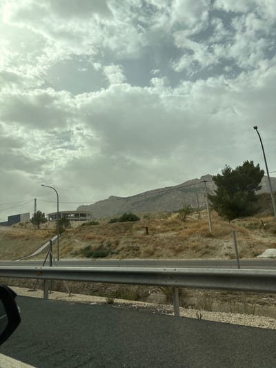 The image depicts a roadside view with a mountainous backdrop under a cloudy sky. The scene is captured from the perspective of someone in a vehicle, as indicated by the reflection of a car's interior in the lower right corner.

Dominating the foreground is a grey asphalt road, bordered by a metal guardrail. Beyond the guardrail, the terrain is dry and arid, covered in sparse, brown vegetation. A tall, dark green tree stands prominently on the hillside, casting a shadow. Further in the distance, a rugged, light-colored mountain range rises against the overcast sky.

Several poles are visible along the roadside, some supporting streetlights or power lines. In the middle ground, there is evidence of construction, with a partially built, circular structure visible. The overall impression of the landscape is one of a dry, somewhat barren environment, likely in a Mediterranean climate.

The sky is filled with dense, grey clouds, suggesting overcast or potentially inclement weather. The lighting is diffused, indicating it is likely daytime, though the specific time is hard to ascertain due to the cloud cover. There is no visible text in the image. The setting appears to be a rural or semi-rural area, possibly along a highway or main road, given the guardrail and the paved surface.