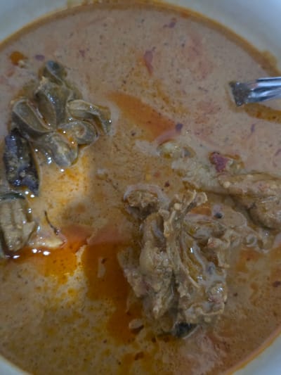 A close-up, overhead shot captures a dish of curry in a white bowl. The curry is a light brown color with visible flecks of red and purple spices. Several pieces of meat, appearing to be chicken or a similar poultry, are submerged in the sauce. The texture of the meat is rough and slightly stringy. A silver spoon is partially visible at the top right of the bowl, suggesting the dish is being served or has been eaten. The lighting is direct and creates a slight sheen on the surface of the curry. The image is focused on the food itself, with no background elements visible other than the rim of the bowl. The scene is likely in a kitchen or dining area in Bireuenmeunasahblang, Indonesia.