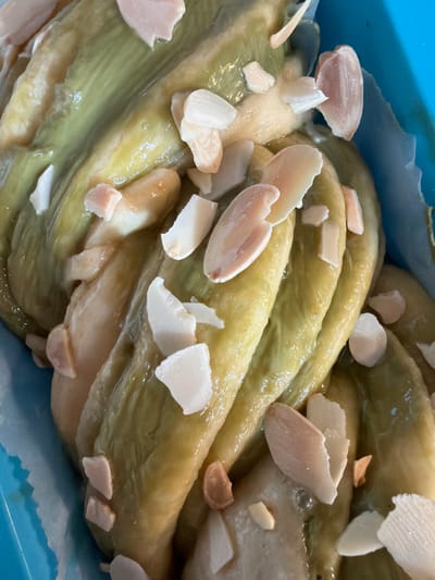 A close-up, high-angle shot displays a green-hued, glossy braided loaf or pastry, likely a dessert. The loaf is arranged in layers or strands, with what appear to be sliced almonds sprinkled on top. The background includes a light blue, possibly a tray or container. The image does not provide information about Bretton, United Kingdom or any people, actions, or interactions in that location.
