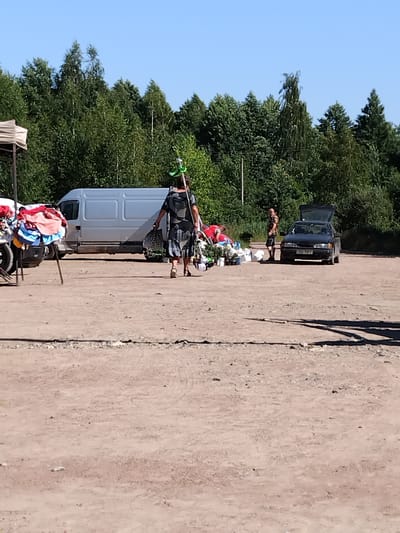A person walks across a dusty lot on a sunny day, carrying a long object with a green attachment over their shoulder and a patterned bag in their other hand. They are dressed in a dark, patterned dress and sandals. Behind them, a light-colored van is parked, and to the right, a dark-colored car with its trunk open is visible. A pile of clothing is draped over a stand on the left side of the image, and another person stands near the car on the right. The background is filled with a dense line of green trees under a clear blue sky. This scene appears to be located in Irshansk, Ukraine.