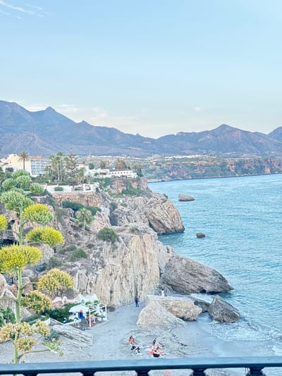 The image captures a scenic view of a rocky coastline leading to a beach, with the turquoise waters of the Mediterranean Sea in the background. In the foreground, lush green vegetation, including striking agave plants with their yellow flower stalks, adds a vibrant touch to the rugged landscape. Several white structures, possibly part of a resort or residential area, are nestled into the hillside, offering a stark contrast to the natural rock formations. The beach below is dotted with a few people sunbathing on loungers and towels, enjoying the sunny weather.

The scene is set in Nerja, Spain, as indicated by the characteristic white architecture and coastal topography. The clear blue sky suggests it is daytime, likely late morning or afternoon, with good weather for beach activities. The composition highlights the natural beauty of the location, with the imposing mountains framing the coastline and the expansive sea. The presence of people indicates a leisurely atmosphere, a typical holiday scene. There are no visible text elements in the image.