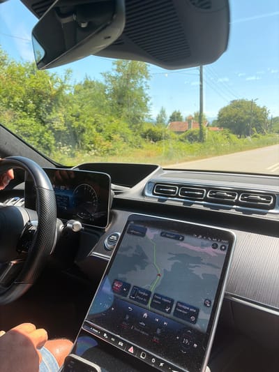 This image shows the interior of a car, likely a Mercedes-Benz S-Class, on a sunny day. The view is from the passenger seat, looking towards the dashboard and the road ahead through the windshield. The car's digital instrument cluster displays speed and other vehicle information, while the central touchscreen shows a navigation map with a route highlighted. The background visible through the windshield suggests a rural or suburban setting with trees, houses with red tile roofs, and power lines. Based on the navigation display which shows the location as "Kaleytsa, Bulgaria", the scene is set in Kaleytsa, Bulgaria. A person's hand and leg are visible in the lower left and bottom center of the frame, respectively, indicating a person is inside the vehicle. The overall impression is of a car journey on a clear day in the Bulgarian countryside.