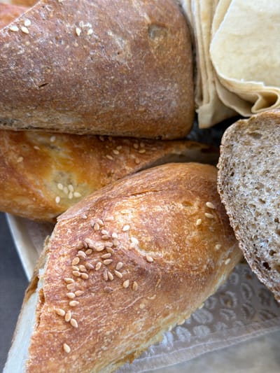 The image shows a close-up of various types of bread. On the left, there is a stack of flatbreads and a loaf of brown bread. In the center, there is a baguette with sesame seeds. A slice of brown bread is on top of the baguette. The bread appears to be in a basket or container. The image was not taken in the city of Lermontovo, Armenia.