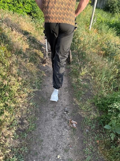 A person is walking on a dirt path in a grassy, overgrown area. The person is wearing black pants and white sneakers. They are pushing a stroller. The environment appears to be rural or semi-rural, with green vegetation and dry grass. The image is taken from behind the person, focusing on their lower body and the stroller.