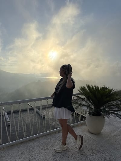 A woman with blonde braids stands on a balcony overlooking a mountainous landscape. The sun shines brightly in the sky, obscured by some clouds. The balcony has a metal railing and is decorated with a large potted plant with lush green fronds. The woman wears a black shirt, white skirt, and white shoes. The location is Kedisan, Indonesia.