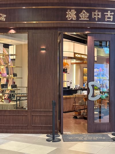 An exterior shot captures the entrance to a store called "MOMO VINTAGE" located in Heping District, China. The store's facade is made of dark wood paneling with gold accents. Above the entrance, Chinese characters are displayed in illuminated gold lettering. The store has large glass windows displaying handbags on shelves. To the right of the entrance is a glass door with a large, stylized "C" shaped handle. Inside the store, wooden shelves are stocked with merchandise. A black retractable stanchion is positioned to the left of the entrance, and a gray welcome mat with the store's name is on the floor in front of the door. The flooring outside the store is tiled in shades of beige and gray.