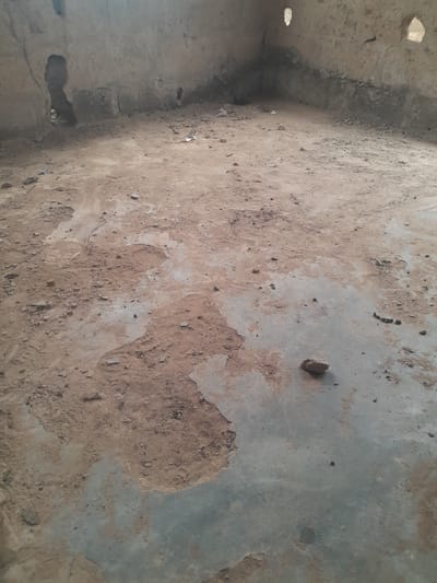 The scene depicts the interior of a severely dilapidated and empty room in Ifo, Nigeria, viewed from a slightly elevated perspective towards a corner. The floor is an uneven mixture of exposed, cracked, and stained concrete or cement, largely covered by a layer of loose, reddish-brown dirt, small stones, and scattered debris. One prominent, irregularly shaped rock or hardened earth clump rests on an exposed concrete patch in the lower-right foreground. The walls, appearing to be made of light-colored, possibly painted or plastered concrete, show significant deterioration with large areas of flaking, missing material, and discolored patches, particularly near the base. Several irregular, dark openings, possibly holes or rough window/door remnants, are visible in the upper sections of the walls. No individuals, furniture, or recognizable personal items are present. The lighting is ambient, suggesting natural light entering from unseen openings or the holes in the walls.