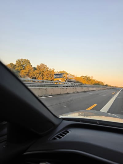 The image, taken from the perspective of a vehicle traveling on a highway, shows a yellow truck with a cylindrical tank on its rear moving in the same direction. The truck has "RENNARD WEISS" written on its side. The scene is illuminated by the warm, low light of sunset, casting long shadows and highlighting the golden hues of the trees lining the highway. The highway itself is asphalt with clear lane markings, including white and yellow lines. A concrete barrier separates the highway lanes from the adjacent foliage. The foreground is dominated by the interior of the vehicle, specifically the dashboard and windshield frame, which are dark and out of focus. The sky is a pale blue, transitioning to a lighter shade near the horizon. The location is identified as Bischofsheim, Germany.