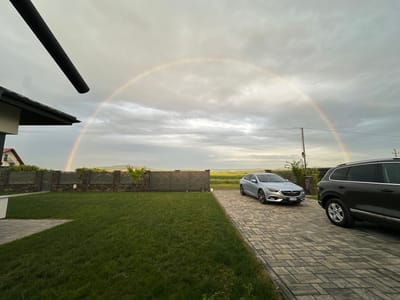 In Târgu Neamț, Romania, a natural phenomenon of a rainbow arches across a cloudy sky, visible above a manicured lawn and a paved driveway. Two cars are parked on the driveway: a silver sedan in the foreground and a dark SUV behind it. The scene also includes a modern fence, a house with a dark roof, and a distant landscape of rolling hills and fields. The lighting suggests it is daytime, possibly after a rain shower, as indicated by the presence of the rainbow.