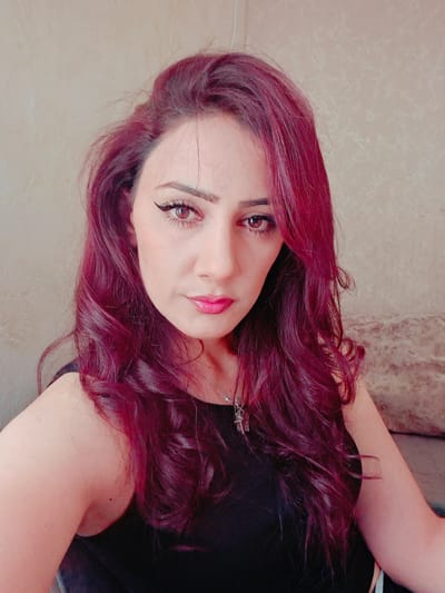 A medium shot shows a woman from the chest up, her head slightly tilted to the right, and her gaze directed towards the camera. Her hair is a dark, wavy, magenta color, falling around her face and shoulders. She is wearing a black tank top, and a silver pendant necklace with a cross charm is visible at her collarbone. Her makeup includes winged eyeliner and bright pink lipstick. Her eyes are a reddish-brown hue. The background is a textured, light-colored wall. The overall lighting is soft, casting a slight shadow on the left side of her face. The image appears to be a selfie.