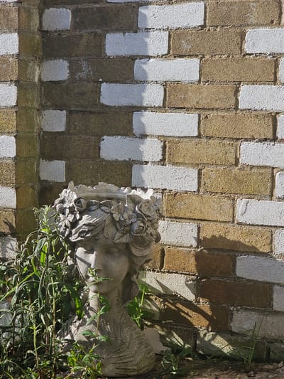 This is an eye-level shot of a brick wall with a stone sculpture resting on top of it. The brick wall is made of alternating rows of tan and white bricks arranged in a staggered pattern. The stone sculpture appears to be a figure with floral embellishments and has some green vegetation growing around it, with some of the foliage obscuring parts of the sculpture. The right side of the image has the edge of a tree trunk. The location might be near some greenery or a garden. Without explicit landmarks or specific architectural details, it is difficult to pinpoint the precise location in Bristol, United Kingdom.