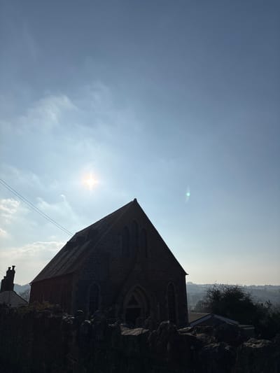 An outdoor, daytime image depicts the Snailbeach Methodist Chapel in Snailbeach, United Kingdom, silhouetted against a bright, hazy sky with partial cloud cover. The primary subject is the dark, stone-built chapel, featuring a steep gabled roof with dormer-like structures on the left slope, and multiple tall, pointed-arch windows. A prominent pointed-arch entrance is centered on the front facade.
The sun, visible as a bright starburst lens flare, is positioned in the upper left quadrant of the sky, casting the chapel and foreground into deep shadow. A smaller, green lens flare appears in the upper right sky. In the immediate foreground, a rough-hewn stone wall, likely dry-stone, runs horizontally across the lower portion of the frame, also heavily shadowed. To the far left, a chimney stack from an adjacent building is partially visible, along with overhead utility wires stretching diagonally across the upper left. The background on the right reveals a distant, hazy landscape of rolling hills and scattered trees. No individuals are present, and no actions or interactions are depicted. The scene is strongly backlit, indicating the sun is positioned largely behind the primary subject.