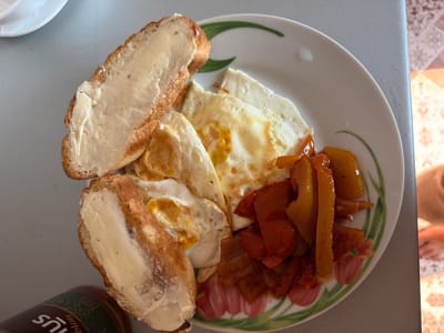 A close-up, overhead shot shows a white plate with a floral design containing a breakfast of fried eggs, sliced bell peppers, and buttered bread. Two slices of bread, generously spread with butter, are placed on the left side of the plate. Three fried eggs with partly runny yolks are arranged in the center, next to a pile of glistening, cooked bell pepper slices in shades of orange and red. The plate sits on a light gray surface, possibly a table. To the bottom left, the neck and part of a dark-colored bottle are visible, with text that includes the word "bus." The background shows a light-colored tiled floor. The image is captured in Norilsk, Russia.