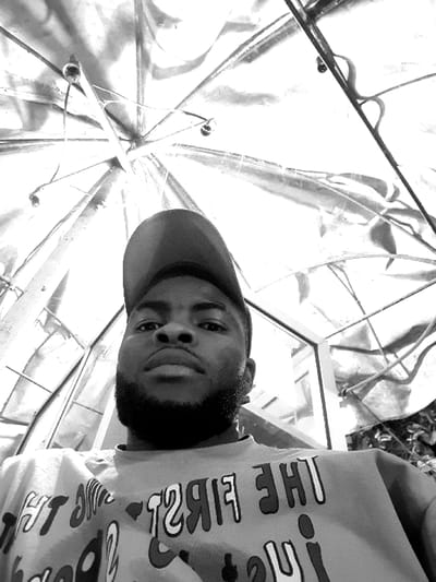 A black and white, low-angle portrait depicts a young adult male with dark skin, a full beard, and a backward baseball cap, looking directly at the camera with a neutral expression. He wears a light-colored t-shirt featuring dark, mirrored text across the chest. The background is dominated by an expansive, reflective overhead structure, resembling an opened metallic umbrella or skeletal canopy, characterized by radial spokes and small, spherical decorative elements. Traces of a glass panel are visible to the subject's left, and dark foliage or plants appear to his right. The scene is situated in Jama A, Nigeria.