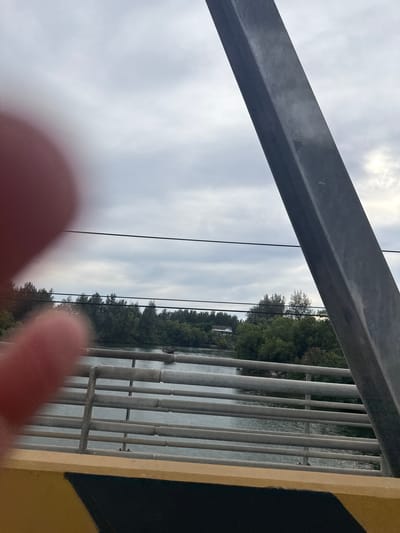 The image captures a view from a bridge in Lamkruet, Indonesia, looking towards a body of water bordered by lush green trees. A large, dark gray metal support beam of the bridge dominates the right side of the frame, extending diagonally upwards against a cloudy sky. In the foreground, a blurred red-pink object, possibly a finger, partially obstructs the view on the left. The bridge railing, composed of horizontal metal bars, runs across the middle ground, with a yellow and black striped barrier visible at the very bottom. The water surface is calm, reflecting the overcast sky and the dense foliage on the opposite bank. A few buildings, one white with a dark roof, are partially visible amidst the trees. Power lines stretch horizontally across the scene in the upper portion. The overall impression is of a natural landscape viewed from an urban infrastructure.
