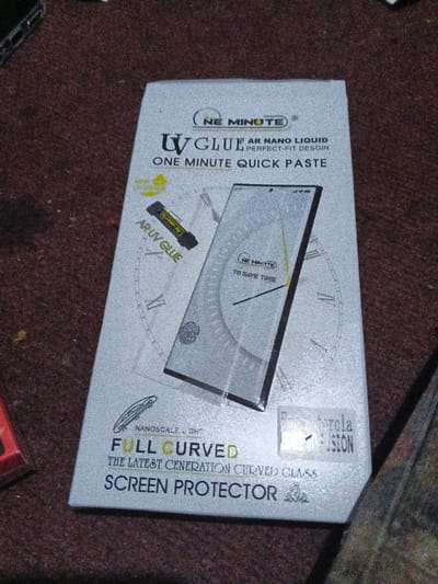 The image shows a box for a "Ne Minute UV Glue" screen protector. It's labeled as "AR Nano Liquid Perfect-Fit Desgin" and "One Minute Quick Paste." The packaging features an illustration of the screen protector on a phone with a clock face behind it. It claims to be "Full Curved" and the "latest generation curved glass" with nanoscale light. There is also a sticker for "Motorola Fusion". An "AR UV GLUE" tube is also shown.