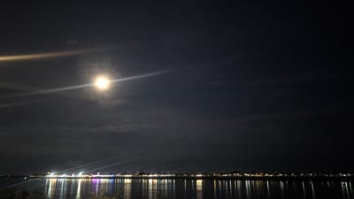 A night-time cityscape is depicted across a wide body of water, likely the Mekong River, with the lights of Vientiane Capital, Laos, stretching along the far bank. The moon, partially obscured by thin clouds, is a bright, luminous orb in the upper left quadrant of the sky, casting a strong glare and lens flare across the scene. The city lights form a continuous, sparkling line along the water's edge, reflecting in the dark, rippling surface of the river. The overall atmosphere is tranquil and illuminated by the ambient light of the moon and city.