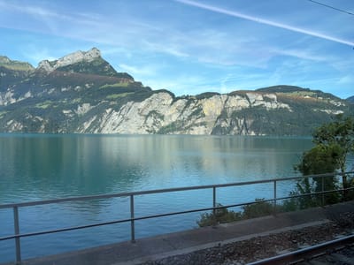 This image captures a scenic view from Morschach, Switzerland. A serene, turquoise lake dominates the foreground, its surface rippled by a gentle breeze. Along the lake's edge, a grey metal railing runs horizontally across the frame, suggesting a walkway or viewing platform. Beyond the railing, lush green trees line the shore, their reflections faintly visible on the water. In the background, imposing mountains rise steeply, their rocky faces contrasted with verdant slopes covered in trees and patches of grass. The sky above is a vibrant blue, streaked with wispy white clouds and a faint contrail from an aircraft. The overall scene conveys a sense of natural tranquility and the majestic beauty of the Swiss landscape.