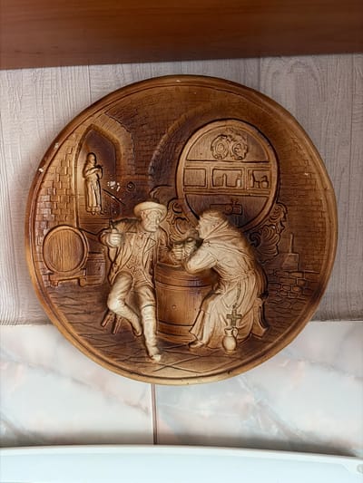 The image shows a circular, bas-relief plaque depicting a scene with two figures and a large barrel. The plaque is made of a brown, wood-like material and appears to be mounted on a wall.

In the foreground, a man in historical attire, including a hat and a long coat, is seated on a stool. He is holding a drinking vessel in his right hand and appears to be gesturing or talking. Beside him, a figure in a monk's robe is kneeling and appears to be interacting with the barrel, possibly tapping it or drawing liquid from it. A rosary with a cross hangs from the monk's belt, and a small jug is placed near his feet.

Behind the figures, a large wooden barrel dominates the background. Above and behind the barrel, there is a depiction of shelves with various vessels. To the left of the barrel, there is another smaller barrel. In the upper left portion of the plaque, a smaller, standing figure in a robe is visible within an arched opening, which suggests a cellar or brewery setting. The background is rendered to resemble brickwork.

The plaque is displayed in what appears to be an interior setting. The wall behind the plaque has a textured, light-colored surface, possibly wallpaper. Above the plaque, a wooden shelf is visible. Below the plaque, there is a surface with a marbled pattern, which could be a countertop or part of a cabinet. The lighting suggests an indoor environment, with a soft light casting subtle shadows on the relief.

There is no indication that this scene is located in the city of Skopin, Russia. The image depicts a decorative plaque and its immediate surroundings, not a specific geographical location within Skopin.