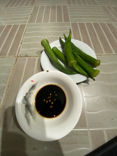 The image shows a plate of okra next to a bowl of dark liquid on a tiled surface. The okra is green and appears to be raw. The bowl contains a dark, glossy liquid, likely a dipping sauce, with small yellow flecks visible within it. The bowl itself is white and has a subtle decorative pattern of grey brushstrokes resembling mountains or clouds, with a small red dot, possibly a stamp, near the rim. The tiled background consists of rectangular tiles in alternating shades of beige and white, with a subtle wave pattern on the white tiles. The lighting suggests it might be indoors, possibly during the day or under artificial light. The overall scene depicts a simple presentation of food, suggesting a meal or snack. No people are visible, and there is no discernible text in the image. The setting appears to be a kitchen or dining area. The image is oriented sideways.