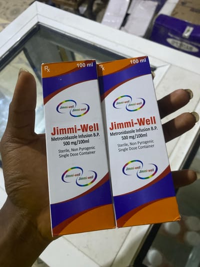 Two boxes of "Jimmi-Well Metronidazole Infusion B.P. 500 mg/100ml" are held in a person's hand. The boxes are white with orange and blue accents. The text on the boxes indicates the product name, strength, and "Sterile, Non Pyrogenic Single Dose Container." The background is blurred, showing a reflective surface that appears to be a tabletop. The image was taken in the city of Jere South, Nigeria.