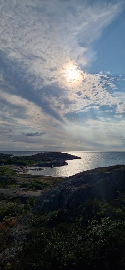 The image depicts a rocky coastline with a small bay and the sea under a cloudy sky. The sun is partially visible, casting a bright reflection on the water. In the bay, there are a few small buildings and a pier. Several people are visible near the water and on the shore, engaged in various activities, possibly swimming or walking. The foreground is dominated by dark, textured rocks and dense green vegetation. The overall impression is of a natural, rugged landscape on a bright but overcast day. This scene is likely from the municipality of Öckerö, Sweden, known for its archipelago and coastal scenery.