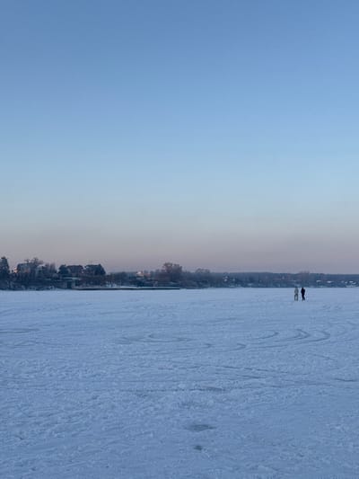 The image depicts a wide, cold-weather landscape in Liutizh, Ukraine, dominated by a vast, frozen body of water. The entire visible surface of the water is covered in a uniform layer of snow and ice, exhibiting numerous faint, curved marks and tracks across its expanse. In the distant background, along the horizon, a shoreline is visible, featuring a mixture of deciduous trees, many appearing bare, interspersed with several residential structures characterized by dark roofs. A low-lying structure, potentially a bridge or embankment, is situated along this shoreline toward the left side of the frame. The sky occupies the upper two-thirds of the image, presenting a gradient from a clear, pale blue at the top to a softer, slightly pinkish hue near the horizon line, indicative of a low sun angle. Two human figures are positioned on the frozen surface in the right midground. The figure on the left wears light-colored clothing, while the figure on the right wears dark-colored clothing. They stand in close proximity, facing generally toward the left, without clear specific actions or interactions discernible beyond their presence.