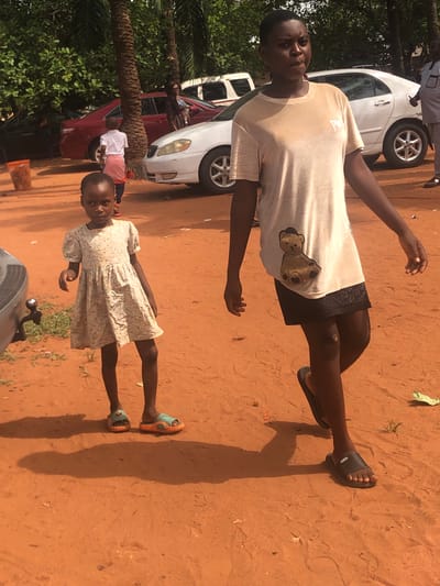 Two individuals are positioned on a dirt surface. The first individual, an adult, is wearing a light-colored t-shirt with a bear image, black shorts, and dark sandals. The second, a child, is wearing a light-colored floral dress and light-colored sandals. The background includes partial views of two cars, one white and one red, along with trees. The location is determined to be Nteje, Nigeria.