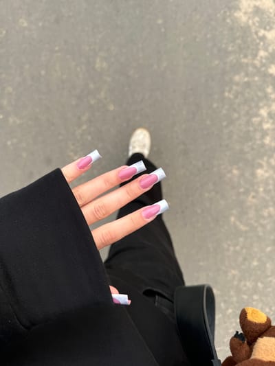 A person's hand with pink and light blue french tip acrylic nails is positioned in the foreground, partially covered by a black garment. The person is wearing black pants and white sneakers, suggesting they are outdoors on a paved surface, possibly a street or sidewalk. A portion of a black bag and a brown plush toy are visible to the right. The background consists of a grey, mottled surface, likely asphalt or concrete. The lighting suggests it is daytime. The image does not provide any specific indicators to determine its location within the city of Сароцкі сельскі Савет, Belarus.