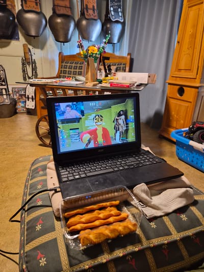 The image shows a close-up of a laptop screen displaying a video game, with a person appearing on a smaller inset screen. The laptop is placed on a patterned cushion, with a white sock and a clear plastic container of food items resting beside it. In the background, there is a wooden cabinet and a window covered by a grey curtain. The overall setting appears to be a room in a home, potentially in Riaz, Switzerland, given the presence of decorative cowbells mounted on the wall, which are often associated with Swiss alpine culture. The scene depicts a person engaged in leisure activity, possibly playing a video game while snacking, in a home environment.